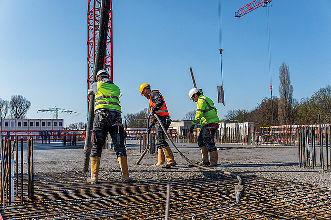 Zwei Bauarbeiter in Schutzkleidung gießen Beton auf eine Bewehrungsschicht. Einer hält den Schlauch zum Betonieren, der andere verteilt den Beton.
