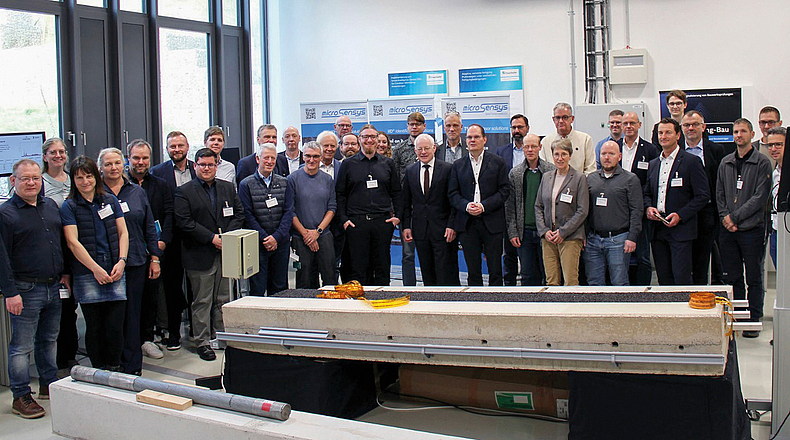 Große Gruppe von Personen steht in einem Laborraum. Im Vordergrund sind technische Geräte und ein Prüfaufbau zu sehen.