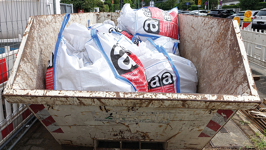Asbesttransport entsprechend der Sondervorschrift 168 Ein Metallcontainer auf einer Baustelle am Straßenrand mit weißen Säcken, die mit Warnhinweisen für asbesthaltige Abfälle gekennzeichnet sind.