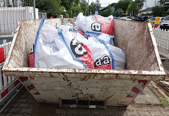 Ein Metallcontainer auf einer Baustelle am Straßenrand mit weißen Säcken, die mit Warnhinweisen für asbesthaltige Abfälle gekennzeichnet sind.