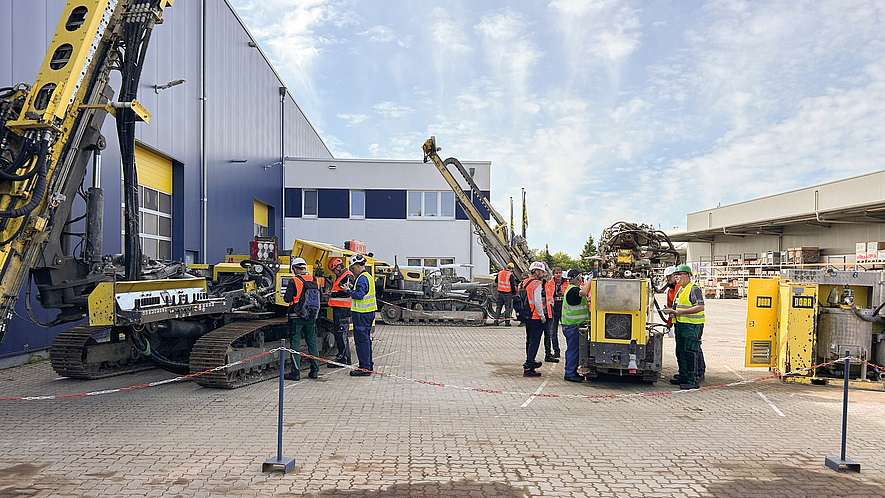 Mehrere Personen mit Warnwesten und Schutzhelmen stehen zwischen Baumaschinen auf einem Betriebsgelände.