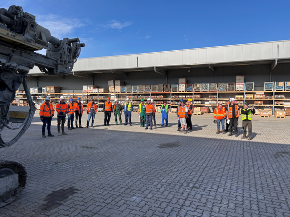 Gruppe von ca. 20 Personen mit orangefarbenen Jacken und weißen Helmen auf einer gepflasterten Fläche. Im Hintergrund ist eine Lagerhalle zu sehen.