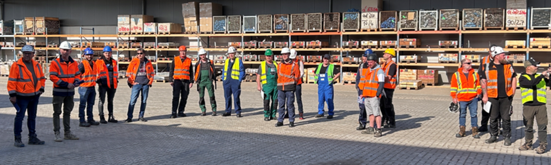 Gruppe von ca. 20 Personen mit orangefarbenen Jacken und weißen Helmen auf einer gepflasterten Fläche. Im Hintergrund ist eine Lagerhalle zu sehen.