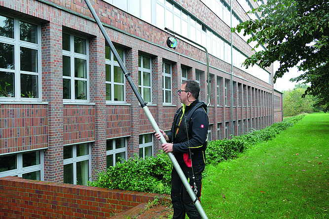 Um die Halswirbelsäule zu entlasten, wird die Teleskopstange in einen Ausleger mit Balancer eingehängt. Ein Mann, der eine Prismenbrille trägt, nutzt eine Teleskopstange mit Ausleger und Balancer sowie Hüftgurt zum Reinigen von Glas und Fassaden.