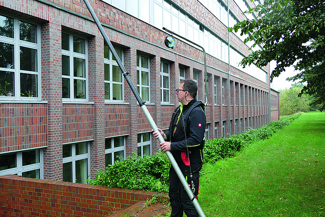 Ein Mann, der eine Prismenbrille trägt, nutzt eine Teleskopstange mit Ausleger und Balancer sowie Hüftgurt zum Reinigen von Glas und Fassaden.