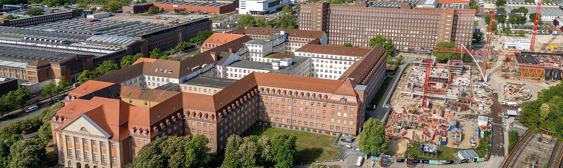Luftaufnahme eines Stadtgebiets mit Baustelle, großen Backsteingebäuden und Industrieanlagen im Hintergrund.