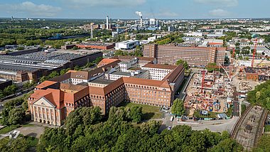 Luftaufnahme eines Stadtgebiets mit Baustelle, großen Backsteingebäuden und Industrieanlagen im Hintergrund.