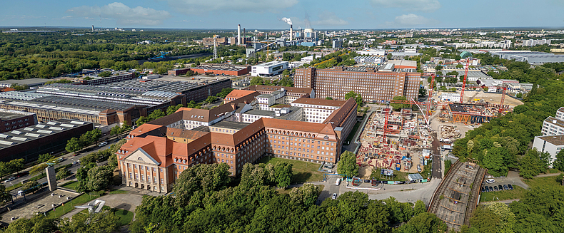 Luftaufnahme eines Stadtgebiets mit Baustelle, großen Backsteingebäuden und Industrieanlagen im Hintergrund.