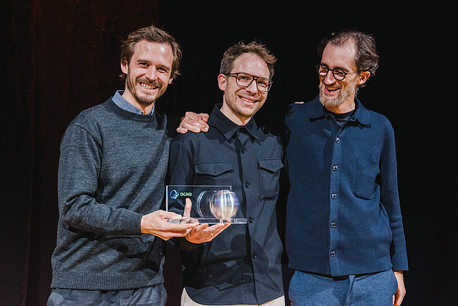 Drei Männer stehen zusammen und halten gemeinsam eine Auszeichnung in den Händen.