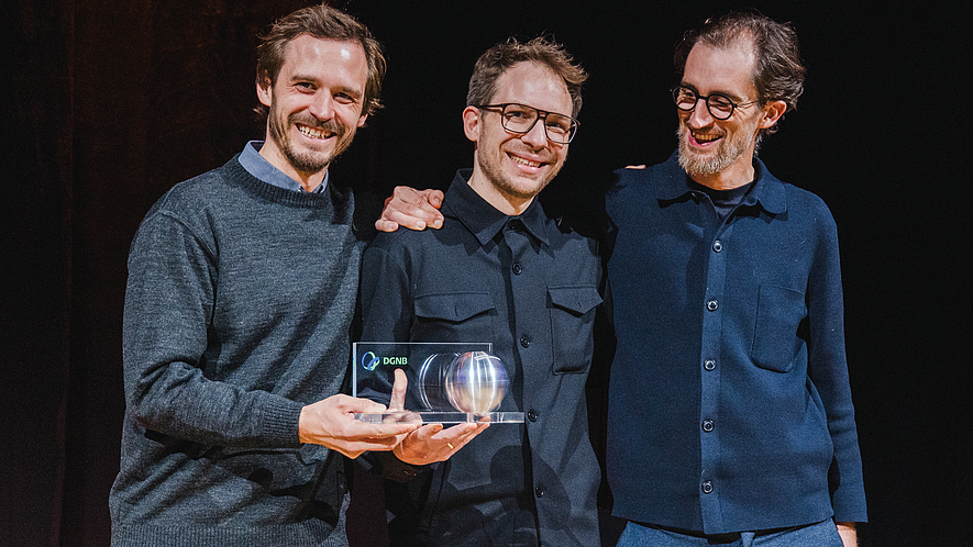 Drei Männer stehen zusammen und halten gemeinsam eine Auszeichnung in den Händen.