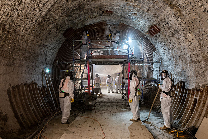 Eine Gruppe von Bauarbeitern entfernt unter Vollschutzkleidung , bestehend aus einem gebläseunterstützten Helm und Schutzanzug, die Fliesenverkleidung der Tunnelinnenschale.
