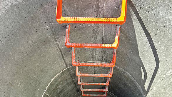 Blick von oben in einen runden Betonschacht mit fest installierten orangenen Steigeisen.