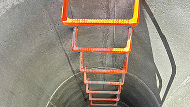 Blick von oben in einen runden Betonschacht mit fest installierten orangenen Steigeisen.