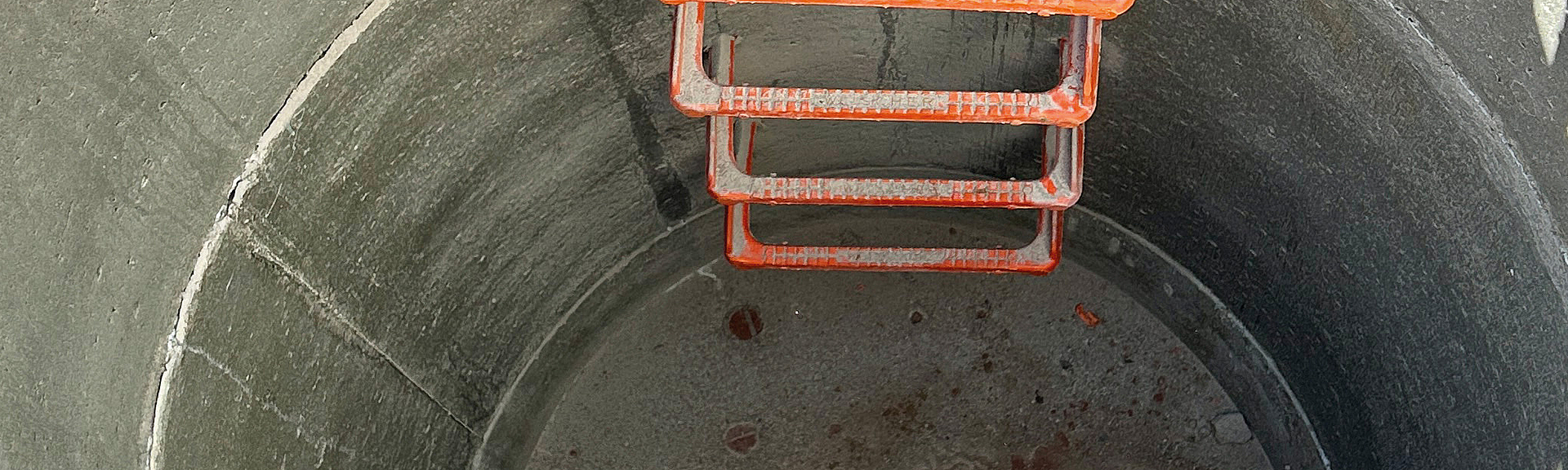 Blick von oben in einen runden Betonschacht mit fest installierten orangenen Steigeisen.