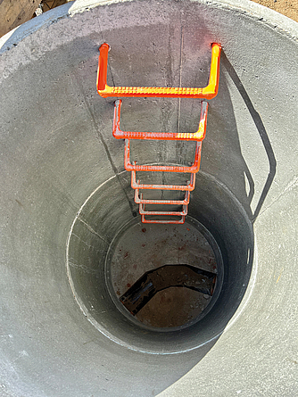 Blick von oben in einen runden Betonschacht mit fest installierten orangenen Steigeisen.