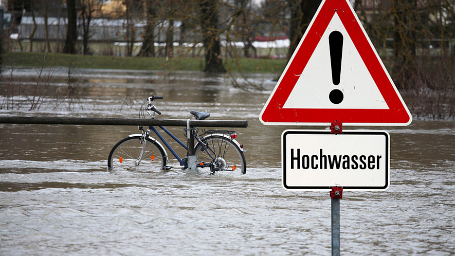 Ein Fahrrad steht im Hochwasser eines Flusses. Warnschild mit der Aufschrift: Hochwasser.