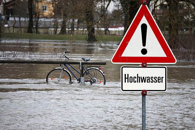 Ein Fahrrad steht im Hochwasser eines Flusses. Warnschild mit der Aufschrift: Hochwasser.