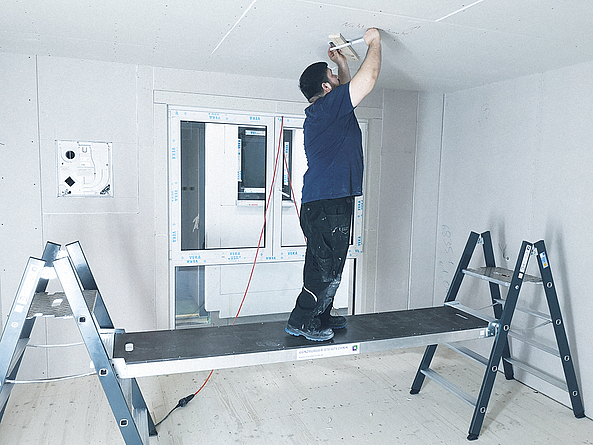 Ein Mann steht auf einer Konstruktion aus zwei Tritten und einer eingehängten Diele und führt über dem Kopf Deckenarbeiten aus.