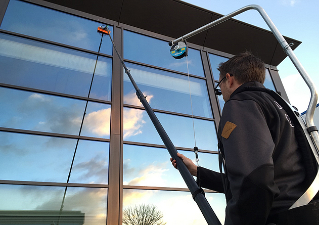 Ein Beschäftigter reinigt Fensterflächen mit einer Teleskopstange mit Bürstenaufsatz, die von einem Rucksacksystem gestützt wird.