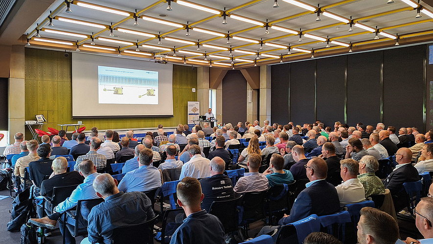 In einem großen Konferenzraum hören zahlreiche Teilnehmende einem Vortrag zu. Auf der Leinwand ist eine Präsentation zu sehen.