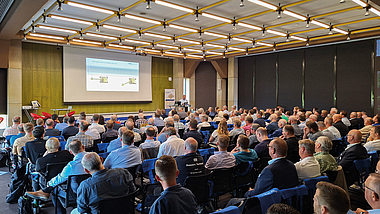 In einem großen Konferenzraum hören zahlreiche Teilnehmende einem Vortrag zu. Auf der Leinwand ist eine Präsentation zu sehen.