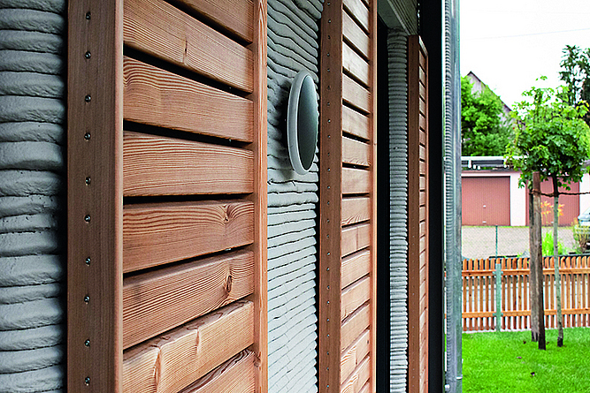 Fassadenausschnitt zeigt den Kontrast zwischen den grauen Wülsten des Betondrucks und den Fensterläden aus braunem Holz 