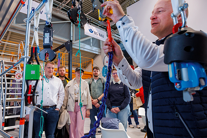 Vorführung zum sicheren Anschlagen von Lastaufnahmemittel und PSAgA am Messestand Ein Mann aus dem Messeteam zeigt einer Personengruppe ein Exponat.