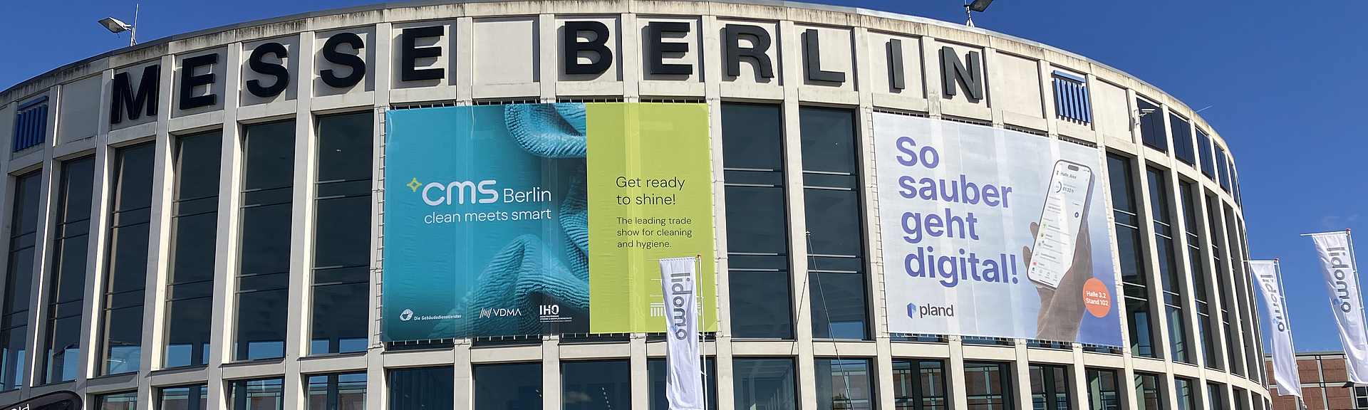 Eingangsgebäude Süd der Messe Berlin, das mit CMS Fahnen behangen ist. Vor dem Gebäude besuchen Gäste die Outdoor-Messestände. 