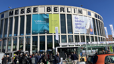 Eingangsgebäude Süd der Messe Berlin, das mit CMS Fahnen behangen ist. Vor dem Gebäude besuchen Gäste die Outdoor-Messestände. 