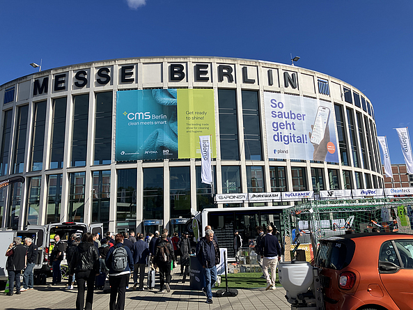 Eingangsgebäude Süd der Messe Berlin, das mit CMS Fahnen behangen ist. Vor dem Gebäude besuchen Gäste die Outdoor-Messestände. 