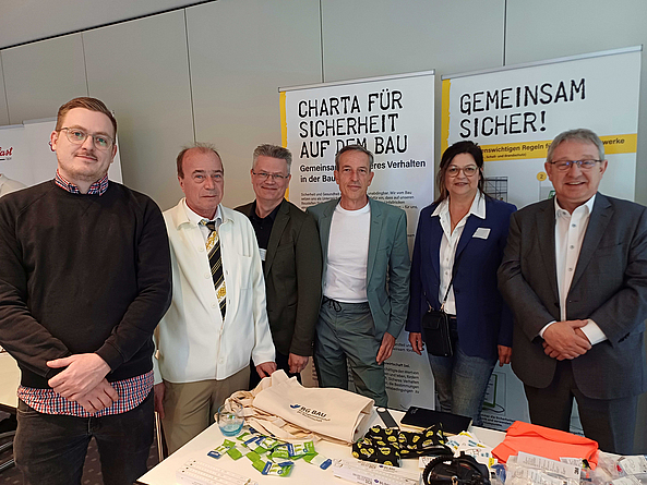 Sechs Personen stehen am Messetisch der BG BAU, hinter ihnen befinden sich große Aufsteller mit der "Charta für Sicherheit am Bau".
