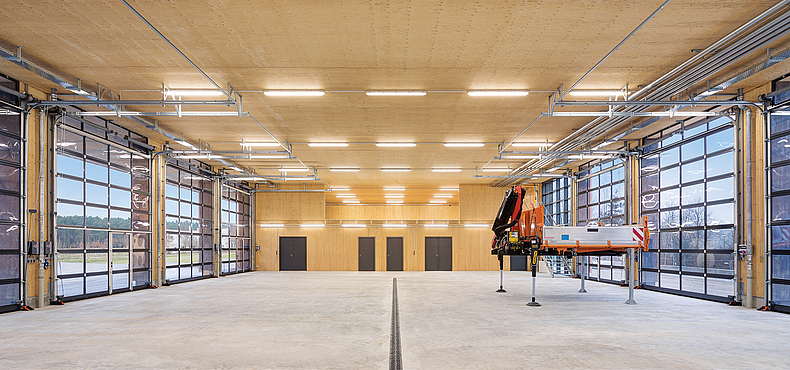 Fahrzeughalle mit angeschalteten Deckenlampen und großen Glastoren, am Rand der Halle befindet sich eine Art Gerüst.