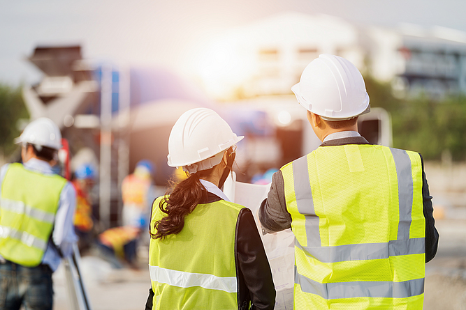 Eine Frau und ein Mann, in Schutzhelm und Warnweste, schauen gemeinsam auf eine Planskizze. Sie befinden sich auf einer Baustelle.