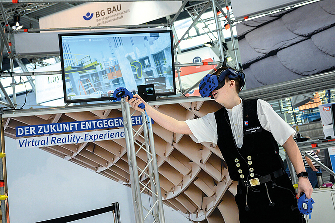 Interaktive Schau der BG BAU: Dank VR-Brille in eine virtuelle Umgebung eintauchen und die Vorteile der Vormontage interaktiv erkunden Mann in Zimmermannskleidung, der an einem Messestand eine VR-Brille trägt und mit dem Scanner etwas erkundet.