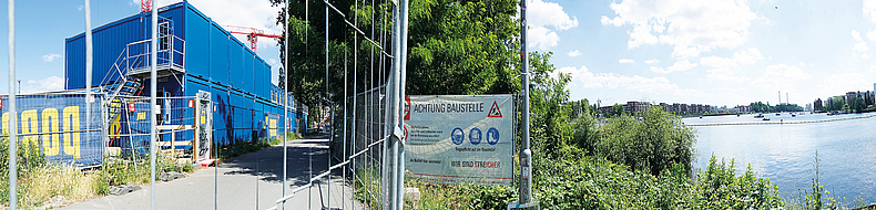 Ein Bauzaun sichert den Uferweg ab. Ein Zaun sperrt einen Weg ab. Hinter dem Zaun ist ein Bauschild.