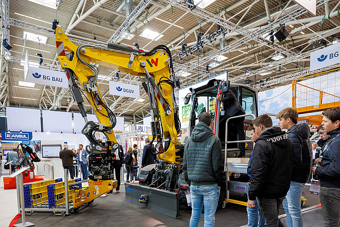 Bagger, der mit einem KI-gestützten Assistenzsystem ausgestattet ist, am bauma-Messestand der BG BAU. Jugendliche betrachten einen Bagger in einer Messehalle, vor dem in einem Gestell gestapelte Module einer Grabenbrücke liegen.