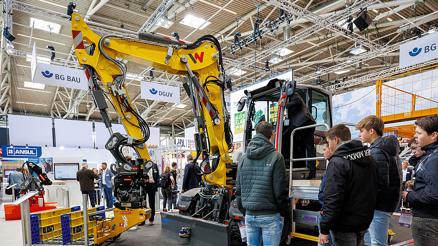 Bagger, der mit einem KI-gestützten Assistenzsystem ausgestattet ist, am bauma-Messestand der BG BAU. Jugendliche betrachten einen Bagger in einer Messehalle, vor dem in einem Gestell gestapelte Module einer Grabenbrücke liegen.