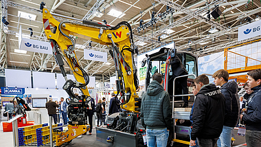 Bagger, der mit einem KI-gestützten Assistenzsystem ausgestattet ist, am bauma-Messestand der BG BAU. Jugendliche betrachten einen Bagger in einer Messehalle, vor dem in einem Gestell gestapelte Module einer Grabenbrücke liegen.