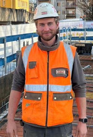 Porträt Timo Holzschuher in Warnweste und Schutzhelm auf einer Baustelle.