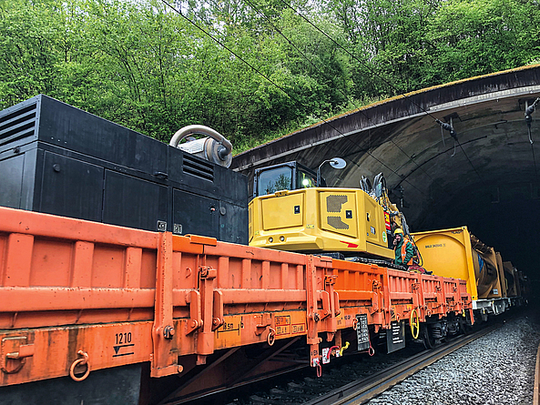 Ein Güterwagen transportiert eine Baumaschine. Daneben steht ein Bauarbeiter mit Schutzhelm und Warnweste.