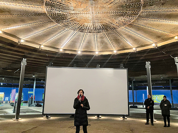 Jeanette Schmitz spricht mit Mikrofon im Foyer des Gasometers. Über ihr hängt eine goldene Gasdruckscheibe.