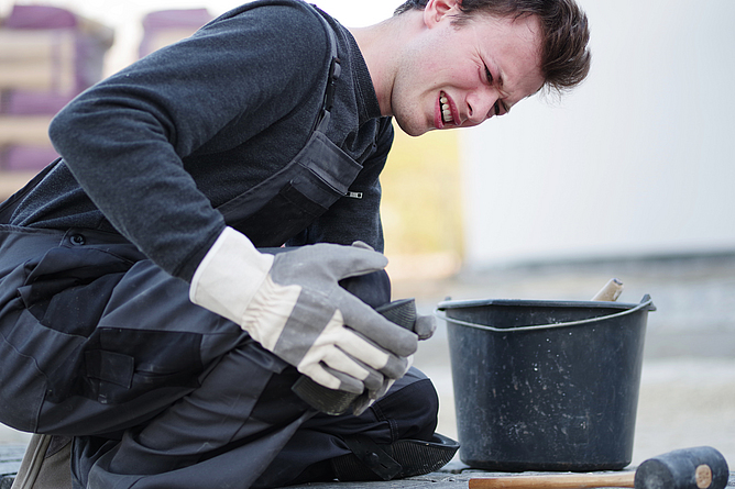Viele Tätigkeiten in der Bauwirtschaft sind belastend für die Knien. Ein Mann mit Knieschmerzen sitzt am Boden und hält sich sein Knie fest.
