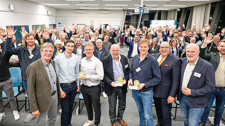 Große Gruppe von Personen steht in einem Veranstaltungsraum, einige halten Auszeichnungen in den Händen.