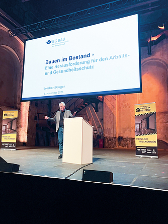 Norbert Kluger sensibilisierte für die Herausforderungen beim Bauen im Bestand. Ein Mann hält auf einer Bühne einen Vortrag. Auf der Leinwand hinter ihm ist der Titel seines Vortrag "Bauen im Bestand. Herausforderung für den Arbeits- und Gesundheitsschutz" zu sehen.