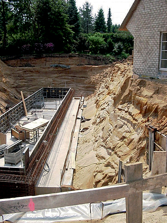 Baugruben aus gleichförmigem (sandigem) Boden können bei entsprechender Witterung und scheinbarer Kohäsion problematisch für die Standsicherheit sein – trotz Böschungswinkel >45°. Längliche Baugrube, an deren rechter Seite eine steile Böschung aus Sand ist.