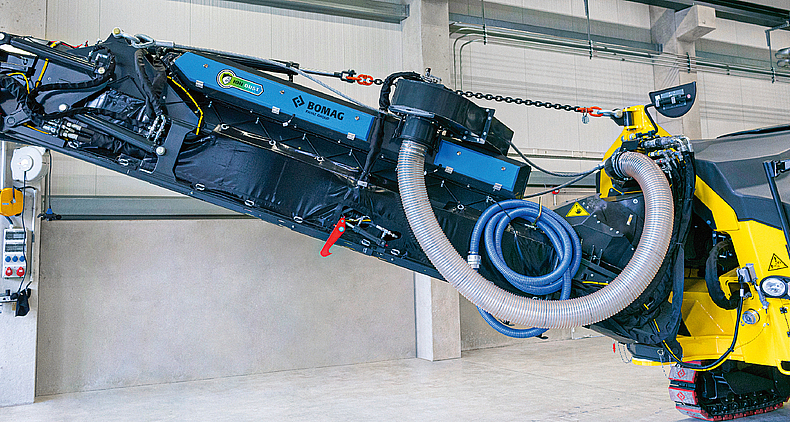 Das ION DUST SHIELD trägt zur Reduzierung der Feinstaubbelastung an Baustellen bei, indem der Staub direkt nach Entstehen beim Fräsprozess abgesaugt und im Gehäuse in Grobstaub umgewandelt wird. Gelbe Straßenfräse, die am vorderen Ende mit einem blauen Elektrostat-Abscheider ausgestattet ist.