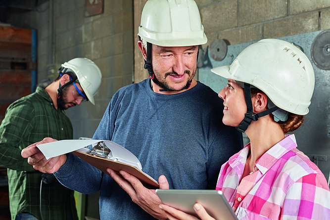 Ein Ausbilder spricht mit einer Auszubildenden auf einer Baustelle. Im Hintergrund arbeitet ein Kollege.