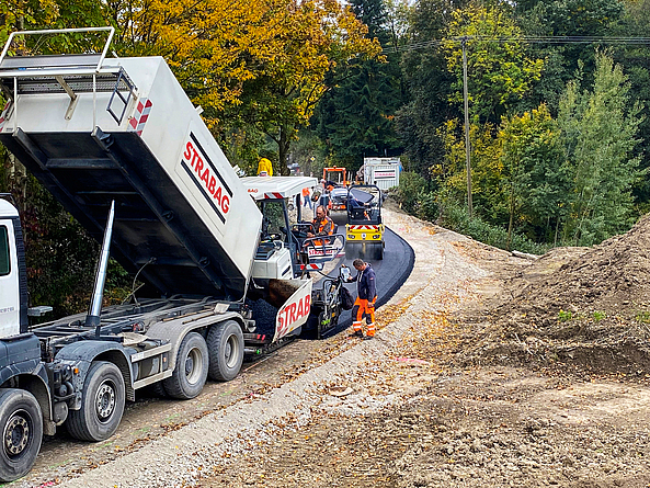 Mehrere Baumaschinen fahren über eine frisch asphaltierte Straße. Vorne am Straßenrand ist ein Bauarbeiter zu sehen.