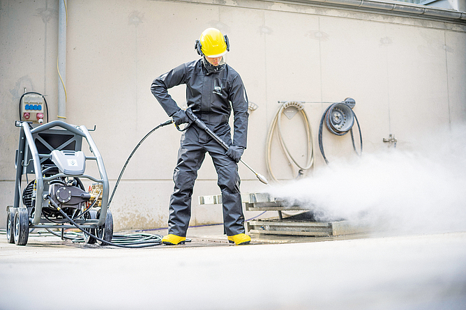 Ein Mann in Schutzkleidung arbeitet mit einer handgehaltenen Spritzeinrichtung und Hochdruckwasserstrahl. Zur Schutzkleidung gehören ein Schutzhelm mit Gesichtsschutz und Gehörschutz, Schutzhandschuhe und Schutzstiefel als geeigneter Fußschutz.
