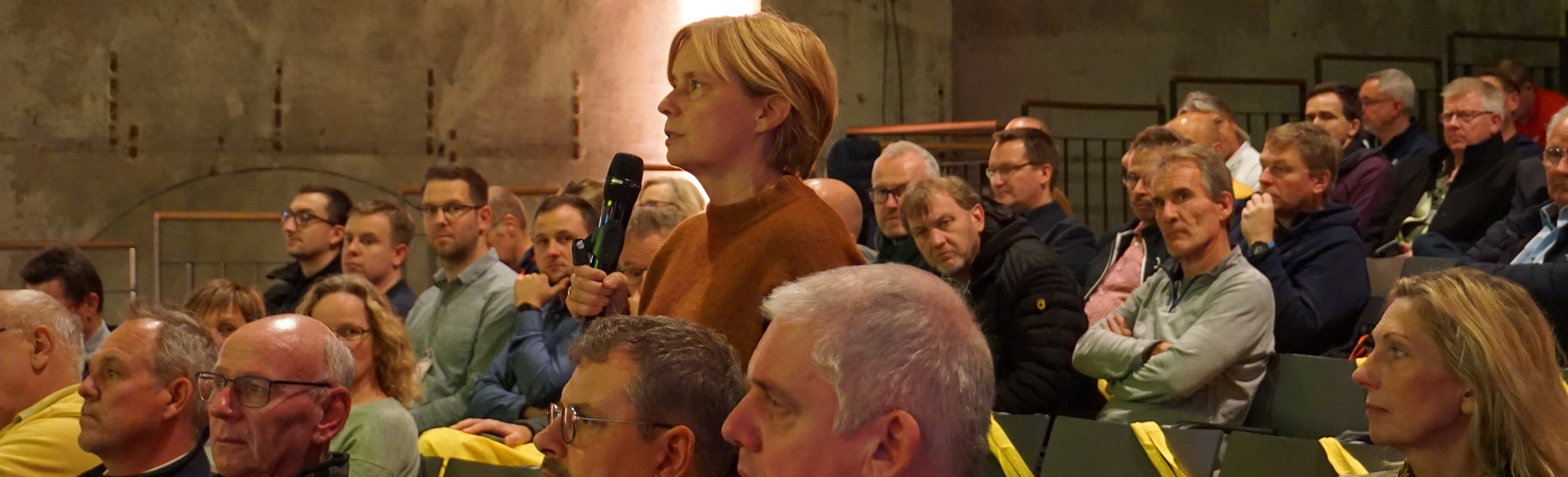 Eine Frau steht mit Mikrofon in einer sitzenden Zuschauermenge und spricht. 